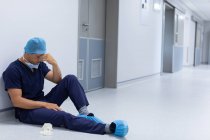 Вид спереди напряжённого кавказского хирурга с лицом на руке, сидящего в коридоре больницы. Хирург носит хирургическую маску, платье, перчатки и кепку . — стоковое фото