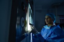 Visão lateral da jovem cirurgiã mista segurando raio-x contra caixa de luz enquanto a examina no centro cirúrgico do hospital. Cirurgião usa vestido cirúrgico, boné e máscara . — Fotografia de Stock