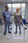 Vista frontal del fisioterapeuta masculino caucásico ayudando a las mujeres caucásicas amputadas a caminar con barras paralelas en el hospital - foto de stock