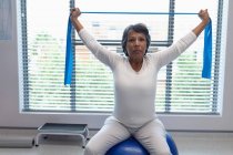 Вид спереду змішаної раси жіночі вправи пацієнта з блакитною смугою опору та м'ячем для вправ у лікарні — стокове фото