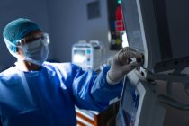 Вид сбоку женщины-хирурга смешанной расы, поворачивающей кнопку хирургического монитора в операционном зале больницы — стоковое фото