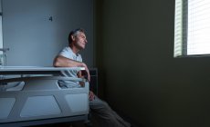 Vue latérale du patient masculin caucasien réfléchi assis sur le lit tout en regardant à l'extérieur de la fenêtre dans la salle à l'hôpital — Photo de stock