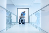 Вид спереду жінка-лікар змішаної раси в хіджабі штовхає старшого пацієнта змішаної раси в інвалідному візку в коридорі в лікарні . — стокове фото