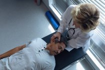Visão de alto ângulo de médico feminino caucasiano examinando mestiço sênior pescoço de paciente masculino no hospital — Fotografia de Stock