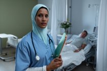 Portrait de belle femme métisse en hijab médecin debout avec rapport médical tandis que le patient masculin caucasien dort en arrière-plan à l'hôpital — Photo de stock