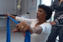 Vista lateral de fisioterapeuta femenina caucásica que da fisioterapia con banda de resistencia a paciente femenina de raza mixta en el hospital - foto de stock
