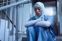 Vue de face de l'infirmière mixte en hijab assise dans les escaliers de l'hôpital — Photo de stock