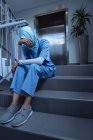 Vista frontal de la triste cirujana de raza mixta en hijab con la mano en la frente sentada en las escaleras en el hospital - foto de stock