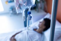Close-up de gotejamento intravenoso com fêmea mestiça deitada na cama em segundo plano na enfermaria do hospital — Fotografia de Stock