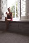 Vorderseite der schönen kaukasischen Frau mit Kaffeebecher sitzt auf Fensterbank im Schlafzimmer zu Hause — Stockfoto