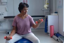 Vista frontal de una hermosa paciente de raza mixta que hace ejercicio con pesas en una pelota de ejercicio en el hospital - foto de stock