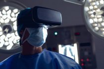 Vista frontal de cirurgiã caucasiana usando fone de ouvido de realidade virtual no centro cirúrgico do hospital. Luzes médicas estão no fundo . — Fotografia de Stock