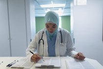 Вид спереду змішаної раси жінка-лікар в хіджабі читання документів на прийомі в лікарні — стокове фото