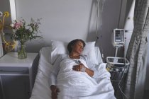 Vista de ángulo alto de la paciente de raza mixta que duerme en la cama con una mano en el estómago en la sala del hospital. Flores están de pie en el armario al lado de la cama . - foto de stock