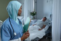 Side view of mixed race female doctor in hijab looking at Caucasian male patient who sleeps in bed in the ward in hospital — Stock Photo