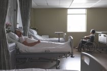 Vue latérale du patient masculin caucasien mature dormant au lit tandis que le patient masculin mixte senior est assis en fauteuil roulant à côté de la fenêtre dans la salle de l'hôpital — Photo de stock