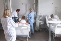 Side view of Caucasian male doctor examining male patient with stethoscope while mixed-race female nurse in hijab is assisting in the ward at hospital. Caucasian female doctor is holding x-ray and senior mixed-race male patient sleeping in bed. — Stock Photo