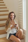 Portrait of happy Caucasian woman with coffee cup sitting on stairs at home — Stock Photo