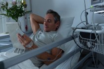 Vue de face du beau patient masculin caucasien utilisant une tablette numérique alors qu'il était allongé au lit avec une main derrière la tête dans la salle à l'hôpital — Photo de stock