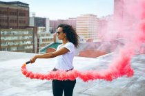 Vue latérale d'une jeune femme afro-américaine portant une chemise blanche et des lunettes tenant un fabricant de fumée produisant de la fumée rouge sur un toit avec vue sur les bâtiments — Photo de stock