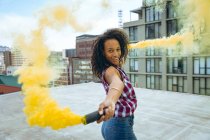 Side view of a young African-American woman wearing a plaid top smiling while holding a smoke maker producing yellow smoke on a rooftop with a view of buildings — Stock Photo