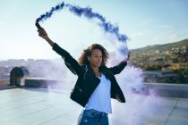 Front view of a young African-American woman wearing a leather jacket holding a smoke maker producing a blue smoke on a rooftop with sunlight — Stock Photo