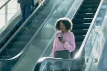 Frontansicht einer afrikanisch-amerikanischen Geschäftsfrau, die im modernen Büro auf ihr Handy schaut, während sie Rolltreppen benutzt — Stockfoto