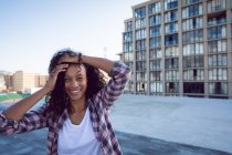 Вид спереди на молодую афроамериканку в клетчатой куртке с руками на голове, улыбающейся на крыше с видом на здание — стоковое фото