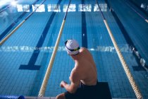 Vista traseira de um nadador caucasiano sentado numa prancha de mergulho junto à piscina — Fotografia de Stock