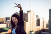 Vista frontale di una giovane donna afro-americana che indossa una giacca di pelle guardando la fotocamera mentre fa un segno di pace e in piedi su un tetto con vista sugli edifici — Foto stock
