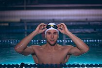 Vista frontal de un nadador caucásico macho sosteniendo las gafas en su gorra de natación blanca mientras está de pie en la piscina - foto de stock