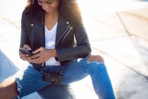 Frontansicht einer jungen afrikanisch-amerikanischen Frau, die eine Lederjacke trägt, eine Kamera am Hals trägt und lächelt, während sie ein Handy benutzt und auf einem Dach sitzt — Stockfoto