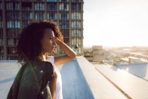 Vista lateral de una joven afroamericana con una chaqueta de cuero sobre el hombro mirando hacia otro lado de la cámara mientras está de pie en una azotea con una vista de un edificio - foto de stock