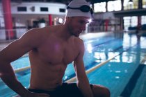 Side view of a male Caucasian swimmer wearing a white swimming cap and goggles sitting by an Olympic sized pool inside a stadium — Stock Photo