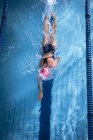 Высокий угол обзора белой женщины в розовой плавательной шапке и очках, делающих фристайл-инсульт в бассейне — стоковое фото