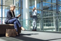 Seitenansicht einer kaukasischen Managerin, die auf ihr Handy schaut, während sie im modernen Büro auf einem Stuhl sitzt. hinter ihr ein afrikanisch-amerikanischer Geschäftsmann, der am Handy spricht. — Stockfoto