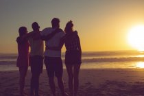 Rear view of diverse friends standing with arms around on the beach during sunset — Stock Photo