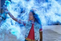 Front view of a young African-American woman wearing a denim vest holding a smoke maker producing blue smoke — Stock Photo