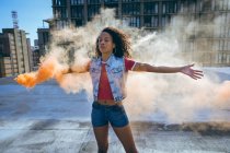 Front view of a young African-American woman wearing a denim vest holding a smoke maker producing orange smoke on a rooftop with a view of a building — Stock Photo