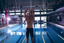 Rear view of a male Caucasian swimmer wearing a white swimming cap stretching by an olympic sized pool inside a stadium — Stock Photo