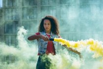 Front view of a young African-American woman wearing a denim vest holding a smoke maker producing yellow smoke on a rooftop with a view of a building and sunlight — Stock Photo