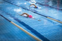 Вид сбоку на молодых афроамериканских и кавказских женщин, делающих фристайл-инсульт в бассейне, в то время как пловец в розовой кепке ведет — стоковое фото
