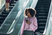 Front view of African american businesswoman looking at mobile phone while using escalators in modern office — Stock Photo