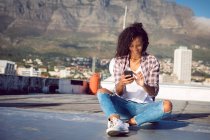 Vista frontale di una giovane donna afro-americana che indossa una giacca a quadri sorridente mentre è seduta e usa un telefono cellulare su un tetto con luce solare — Foto stock