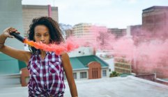 Frontansicht einer jungen afrikanisch-amerikanischen Frau, die ein kariertes Oberteil trägt, während sie eine Rauchmaschine hält, die roten Rauch auf einem Dach erzeugt, mit Blick auf Gebäude, während sie aufmerksam in die Kamera blickt — Stockfoto