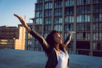 Side view of a young African-American woman wearing a leather jacket looking away from the camera with arms raised while standing on a rooftop with a view of a building — Stock Photo