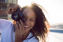 Vista frontale di una giovane donna afro-americana sorridente mentre tiene in mano una macchina fotografica e in piedi su un tetto con vista su un edificio e la luce del sole — Foto stock