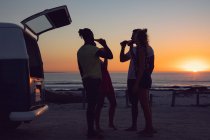 Vista lateral do grupo de diversos amigos bebendo cerveja perto de van campista durante o pôr do sol — Fotografia de Stock
