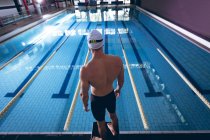 Rückansicht eines männlichen kaukasischen Schwimmers mit weißer Schwimmmütze, der an einem olympischen Pool in einem Stadion steht — Stockfoto