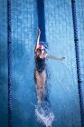 Высокий угол обзора белой женщины в розовой плавательной шапке и очках, делающих удар сзади в бассейне — стоковое фото